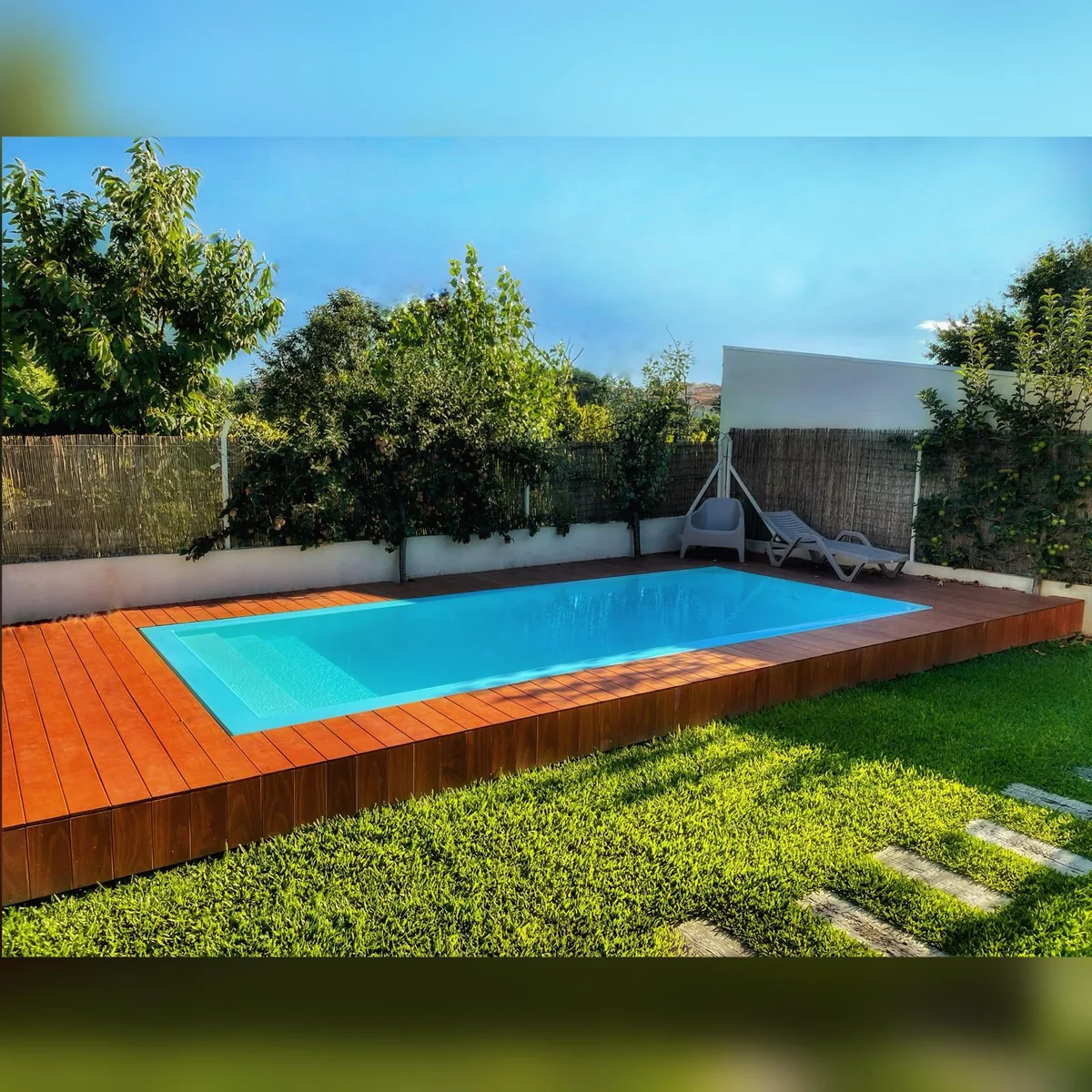 Piscina azul integrada num deck de tom madeira, rodeada de árvores e relva